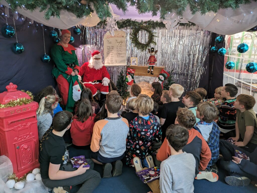 Photo showing Horsmonden pupils sat on the floor in Santa's Grotto, listening to him speak to them.