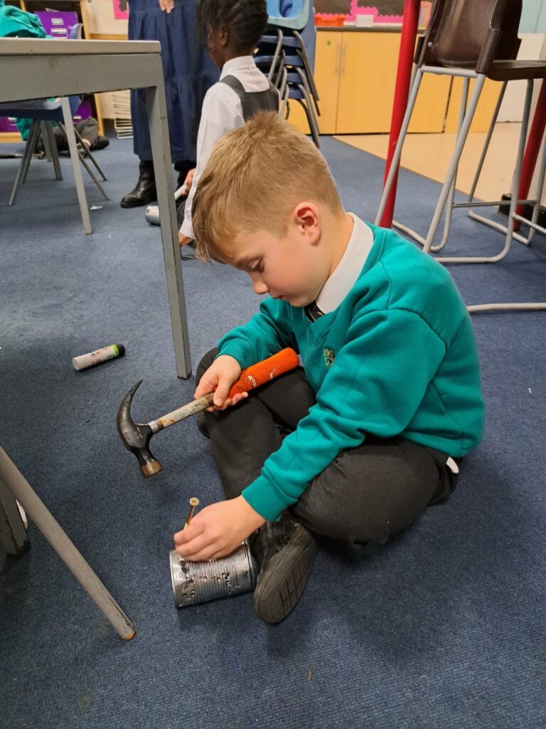 A young boy in academy uniform is pictured holding a hammer in one hand and using it to put a nail into a tin can.